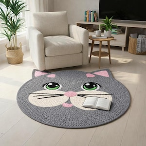 May include: A gray, round cat-shaped rug with a smiling face, pink accents, and green eyes. The rug has a white muzzle and whiskers. An open book rests on the rug. The rug is on a light wood floor.