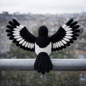 May include: A crocheted magpie with open wings, showcasing a black body, wings, and tail, with white detailing on the wings and chest. The bird is positioned on a metal bar, set against a blurred urban backdrop.
