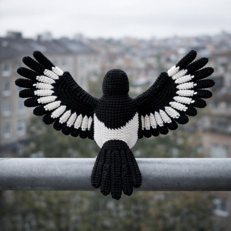 May include: A crocheted magpie with open wings, showcasing a black body, wings, and tail, with white detailing on the wings and chest. The bird is positioned on a metal bar, set against a blurred urban backdrop.