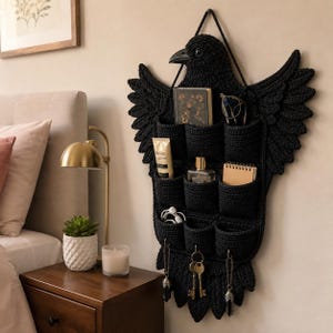 Op de afbeelding: Een zwarte gehaakte wandorganizer in de vorm van een vogel met gespreide vleugels. Het item heeft negen individuele opbergvakken en drie metalen haken aan de onderkant voor het ophangen van voorwerpen, gemaakt van donker getextureerd garen.