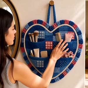 May include: A heart-shaped, blue crochet wall organizer with pockets, holding makeup brushes, a comb, and beauty products. The organizer has a patchwork design with red and white accents, and a blue rope for hanging.