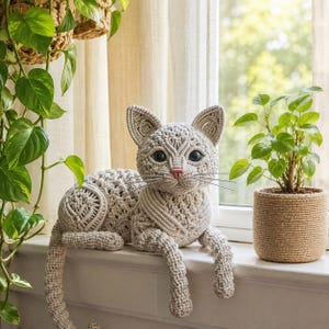 Può includere: Una statuetta di gatto in macramè color beige chiaro con intricati motivi a corda annodata, grandi occhi scuri, un piccolo naso rosa e sottili baffi di filo metallico. Il gatto è realizzato con una trama dettagliata su corpo, orecchie e coda.