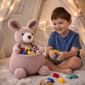 Puede incluir: Una cesta de juguetes de conejo rosa hecha a mano con ganchillo, llena de coches y bloques de juguete de colores. El conejo tiene orejas grandes, una cara blanca y ojos negros. Un niño pequeño está sentado cerca, jugando con bloques. La cesta mide unos 30 cm de alto.
