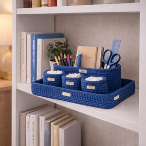 Puede incluir: Un juego de organizador de escritorio de ganchillo azul con una bandeja, un soporte rectangular grande y tres soportes redondos más pequeños. El juego contiene bolígrafos, lápices, tijeras y una regla. Los soportes más pequeños contienen objetos blancos.