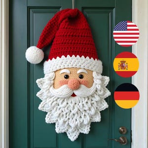 Op de afbeelding: Een gehaakte kerstman-wandhanger met een felrode muts met een witte pompon, een perzikkleurig gezicht, zwarte ogen, een ronde neus en een dikke, getextureerde witte baard, opgehangen aan een donkergroene deur.