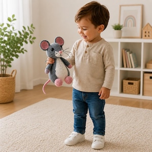 May include: A handmade crochet mouse toy in gray, white, and pink. The plush toy has a long pink tail, pink ears, and pink feet. The mouse is being held by a child wearing a beige long-sleeve shirt and blue jeans.