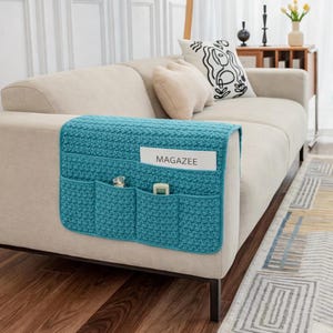 Op de afbeelding: Een turquoise gehaakte armleuning organizer voor een bank met meerdere vakken. De organizer bevat een tijdschrift, een kleine parfumfles en een afstandsbediening. Het woord "MAGAZEE" is zichtbaar op het tijdschrift. De organizer is geplaatst op een beige bank.