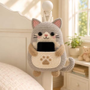 Puede incluir: Un organizador con forma de gato tejido en gris y blanco con un bolsillo para un teléfono. El gato tiene orejas internas rosas, ojos negros y una huella de pata marrón claro en el bolsillo. El organizador cuelga de un marco de cama blanco.
