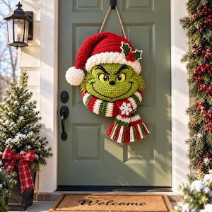 May include: A crocheted wall hanging featuring a green character face wearing a red and white Santa hat with a pom-pom, holly leaves, and berries. The character wears a striped red, white, and green scarf with a white snowflake applique.