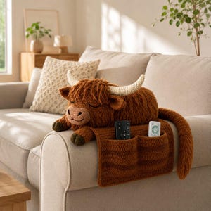 May include: A brown crocheted Highland cow armrest organizer featuring two pockets, cream-colored horns, and a long tail. The organizer is draped over a sofa arm, holding a black remote control and a white remote control in its front pockets.