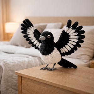 May include: A handmade crochet magpie bird with outstretched wings, showcasing black and white plumage. The bird has a white breast and a grey beak, perched on a wooden surface. The wingspan is approximately 25 cm.
