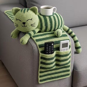 May include: A green and dark green striped crochet cat-shaped sofa armrest organizer. The cat has a smiling face and holds a white mug. Two pockets hold a remote control and a thermostat.