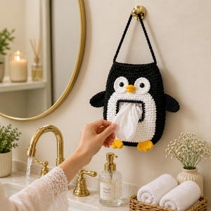 May include: A black and white crochet penguin tissue holder with yellow accents. The penguin-shaped holder is hanging on a wall, with a hand pulling out a tissue. The holder has a black strap and is in a bathroom setting.