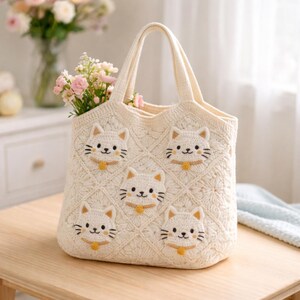 May include: Cream-colored crochet tote bag with five cat faces. The bag has a diamond pattern and a handle. The cat faces are white with black whiskers, eyes, and a yellow collar. The bag is filled with pink flowers.