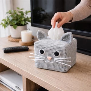 Può includere: Un copri scatola di fazzoletti grigio all'uncinetto a forma di gatto. Ha occhi neri, un naso rosa e baffi bianchi. Anche le orecchie del gatto sono grigie. Una mano estrae un fazzoletto dalla parte superiore. La scatola dei fazzoletti è su una superficie di legno.