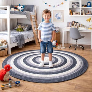 May include: A round rug with concentric circles in shades of gray and white. The rug is on a wooden floor in a child's room. A young child is standing on the rug. The room has a bunk bed, desk, and toys.