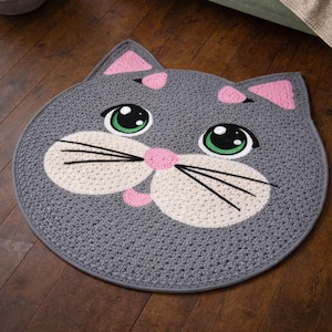 May include: A gray, cat-shaped rug with a crocheted design. The rug features a cat face with large green eyes, pink accents, and black whiskers. The rug is round and made of a soft material.