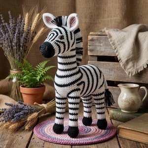 Może przedstawiać: Ręcznie robiona szydełkowa zebra z czarno-białymi paskami, stojąca na okrągłym, wielokolorowym dywanie. Zebra ma czarną grzywę, ogon i kopyta. Tło obejmuje rośliny, drewnianą skrzynkę i ceramiczny dzbanek.