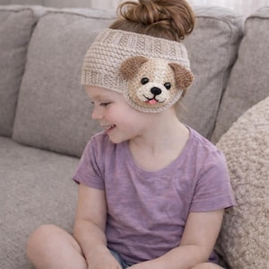 May include: A beige crocheted headband featuring a dog face design. The dog has brown ears, a beige muzzle, black eyes, and a pink tongue. The headband is worn by a child, and the background is a neutral gray.