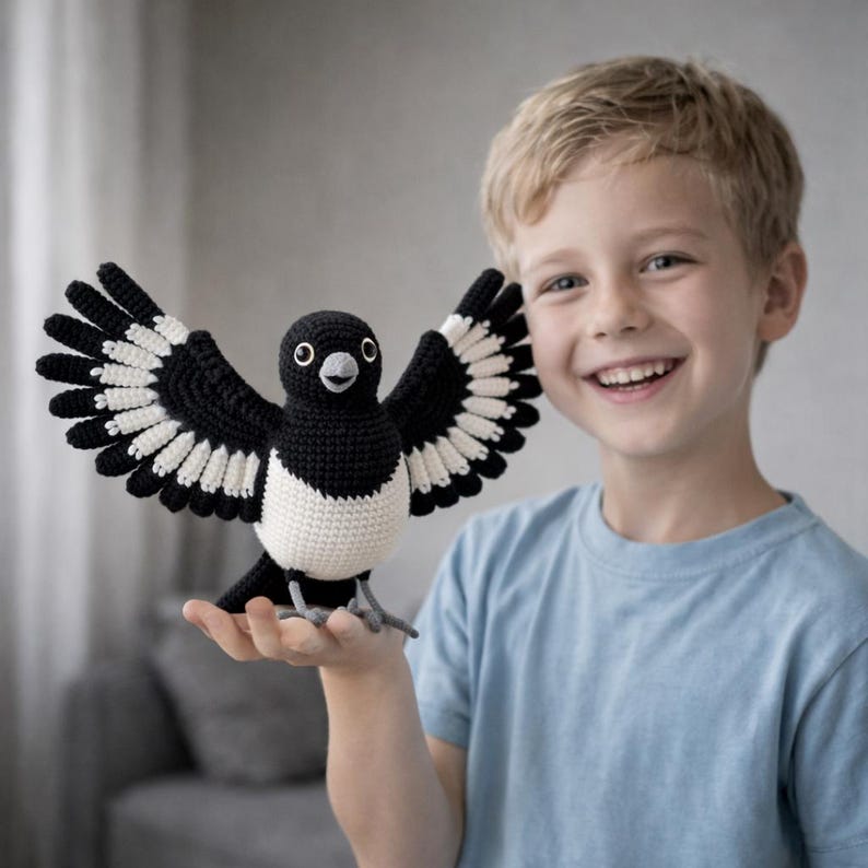 May include: A crocheted magpie bird with outstretched wings, in black and white. The bird has a white belly and is held in a person's hand. The bird's wings are spread wide, showcasing the detailed crochet work. The bird has a friendly expression.