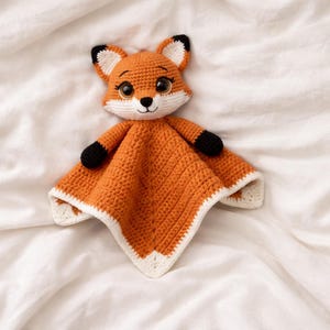 K&ouml;nnte beinhalten: Eine geh&auml;kelte orangefarbene Baby-Sicherheitsdecke in Form eines Fuchses. Der Fuchs hat ein wei&szlig;es Gesicht, Ohren und eine wei&szlig; umrandete Decke. Die Ohren des Fuchses haben schwarze Spitzen und die Pfoten sind schwarz. Die Decke liegt auf einer wei&szlig;en Oberfl&auml;che.