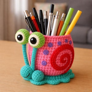 May include: A colourful, handmade crochet pen pot in the shape of a snail. The pink body has a red spiral and purple dots. The snail has green eyes and a turquoise base, holding pens and markers.