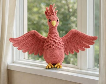 May include: A handmade, coral-coloured crochet bird with outstretched wings. The bird has a yellow beak and feet, and a textured body. The bird is sitting on a white windowsill, with a blurred green background.