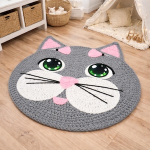 May include: A gray, crocheted cat-shaped rug with pink accents on the ears and nose. The rug features large, white eyes with green irises and black whiskers. This decorative rug is designed for a child's room.