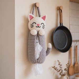 May include: A crocheted cat-shaped plastic bag dispenser in cream and gray yarn. The cat head features pink inner ears, embroidered black eyes, whiskers, and a pink nose. The gray body has a central opening for storing bags and a tail.