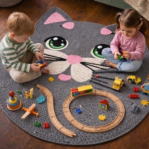 May include: A gray, cat-shaped rug with pink accents and green eyes. Children are playing with colorful toy cars, a wooden train set, and other toys on the rug. The rug is round and made of a soft material.