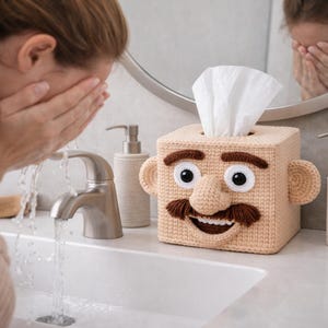 May include: A beige crocheted tissue box cover shaped like a face with large eyes, a nose, a mustache, and ears. White tissues are visible at the top. The box is on a bathroom counter.