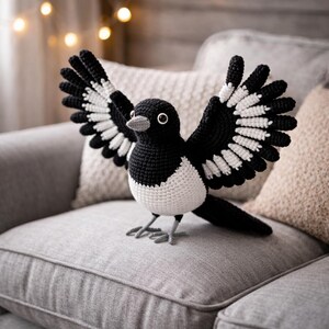 May include: A crocheted magpie bird with open wings, in black and white. The bird has a white chest, black wings with white detailing, and a grey beak and feet. It is sitting on a grey sofa, a decorative item.
