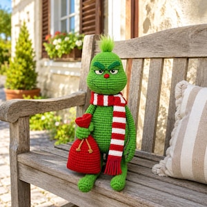 Op de afbeelding: Een groene gehaakte pop met een pluk haar op het hoofd, een rood-wit gestreepte sjaal en een rode zak in één hand. De pop heeft een norse gezichtsuitdrukking met donkergroene wenkbrauwen, een kleine rode neus en een gestikte mond.