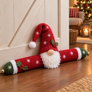 May include: A red and green crocheted draft stopper featuring a gnome with a white beard, a tall red hat with white snowflake embroidery, and small holly leaf accents. The ends of the long cylindrical tube are finished with white bands and gold bells.