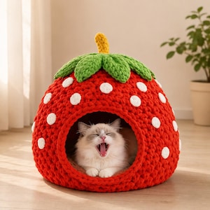 May include: A red crocheted cat bed in the shape of a strawberry, featuring white spots, green leaves, and a yellow stem. A white and grey cat yawns inside the bed. This cat bed is a cosy and decorative pet accessory.