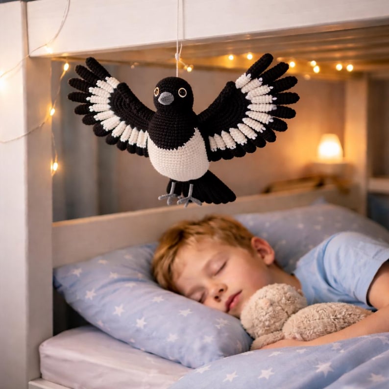 May include: A crocheted magpie, in black, white, and grey, with wings spread, hangs above a child sleeping in a bed. The bed has a blue pillowcase with a star pattern. Fairy lights are strung above the bed, creating a soft, warm glow.