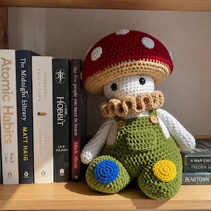 May include: A crocheted mushroom doll with a red cap featuring white spots, a white head, and a tan ruffled collar. The doll wears olive green overalls with two wooden buttons and has a blue circular patch on one knee and a yellow one on the other.