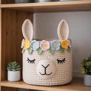 Könnte beinhalten: Ein beigefarbener Häkel-Aufbewahrungskorb in Lama-Form mit großen Ohren und einem Blumenkranz. Der Korb hat ein lächelndes Gesicht mit Wimpern und eine dekorative Blumengirlande. Der Korb steht auf einem Holzregal.
