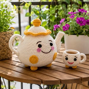 Può includere: Una teiera all'uncinetto color crema con coperchio, manico e base gialli, dotata di occhi a bottone neri, guance rosa e un sorriso ricamato. Accanto si trova una piccola tazza all'uncinetto abbinata con occhi a bottone neri e un sorriso rosso.