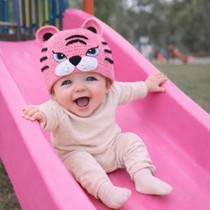 May include: A baby wearing a pink tiger-themed knit hat with black stripes and a white muzzle, smiling widely while sliding down a pink slide. The baby is wearing a beige outfit.