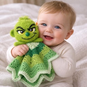 May include: A smiling baby holds a green crocheted security blanket and a matching Grinch-faced doll. The blanket features various shades of green in a square pattern. The doll has black eyebrows, yellow eyes, and a green nose.