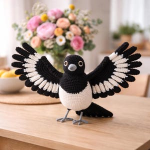 May include: A crocheted magpie bird with open wings, crafted in black and white yarn. The bird has a black head, wings, and tail, with a white chest and wing detailing. It is positioned on a wooden table, showcasing its handmade quality.
