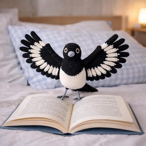 May include: A crocheted magpie with black and white plumage, wings spread wide, standing on an open book. The bird's body is white, with black wings and a black head. The book has light pages and a blue cover.