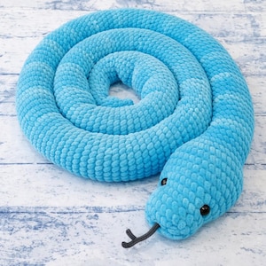May include: A coiled, plush, light blue snake toy. The snake has black button eyes and a dark gray forked tongue. The body is made of a soft, textured material, and the snake is resting on a light-colored wooden surface.