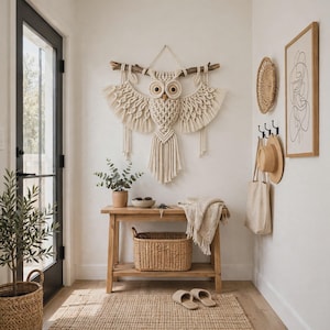 May include: A cream-colored macrame owl wall hanging suspended from a wooden branch. The owl features intricate knotted patterns, large circular eyes, and long fringe wings. It is displayed on a white wall above a wooden console table.