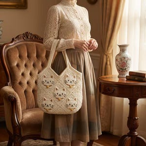 May include: Cream-colored crochet tote bag featuring a repeating pattern of cat faces with yellow bow ties. The bag has two handles and a diamond-shaped stitch pattern. The cat faces have black embroidered details. The bag is held in front of a person.