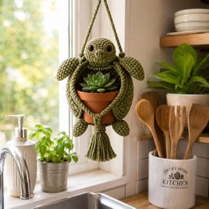 May include: A crocheted olive green turtle-shaped plant hanger holding a small terracotta pot with a green succulent. The turtle has large dark eyes and a textured shell pattern, suspended by a matching cord with a decorative tassel at the base.