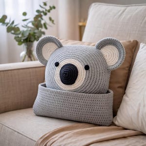May include: A handmade gray crochet koala bear pillow with a matching basket. The koala has a dark blue center, white ring, and black eyes. The ears are gray with white centers. The pillow is sitting on a beige couch.