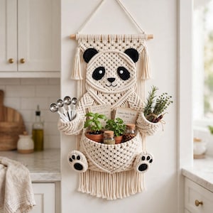 May include: A cream-colored macrame wall hanging shaped like a panda bear, featuring black accents on the ears, eyes, and paws. The panda has two side pockets holding metal measuring spoons and herbs, and a large central pouch for storage.