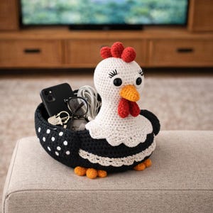 May include: A handcrafted crochet chicken basket in black and white, with a red comb and wattle, and orange feet. The basket contains a black smartphone, glasses, keys, and a white cable. The chicken basket is a decorative storage solution.